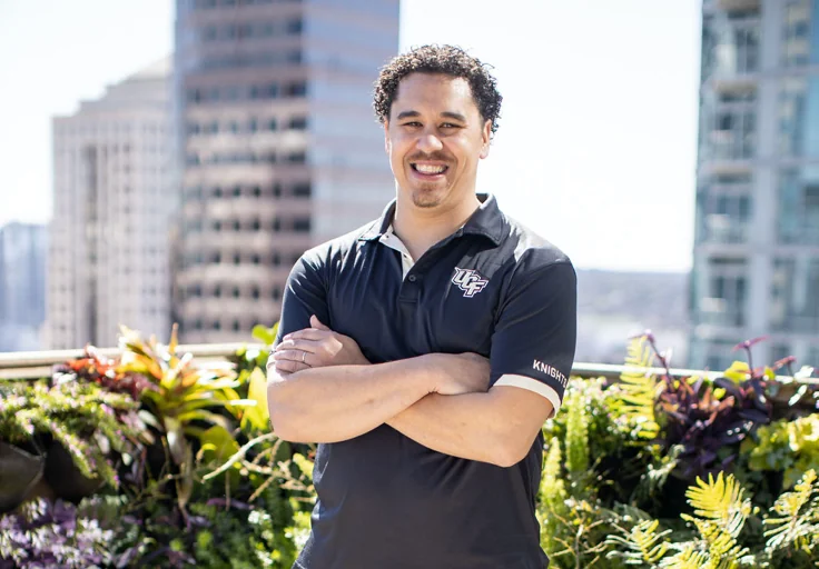 John smiling with his arms crossed in a UCF polo, with greenery and city buildings behind him.
